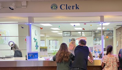 A customer service window with the word Clerk above it. A man and a woman being helped by a female employee. Other people waiting by the side. A woman behind window off to the side working.