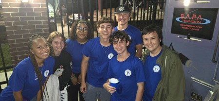 Group of 7 teens wearing Bloomfield Township logo t-shirts standing outside by a tv production truck in the evening.