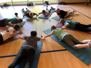 people in a gym lying face down on a yoga mat. They are all facing inward to form a circle and holding hands with arms raised in a skydiver pose.