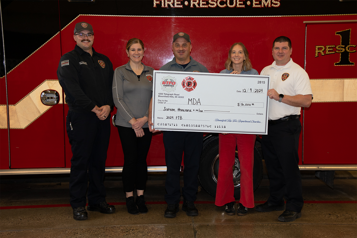 Three men and two women standing in front of a fire truck and holding a large check. 