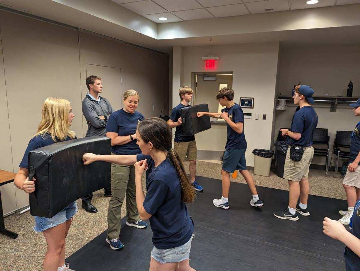 A woman and a man leading a group of teenagers in learning how to punch a block. 